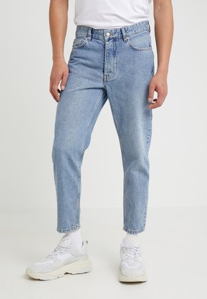 Mann trägt hellblaue High-Waist-Jeans, weiße Turnschuhe und weiße Socken vor einfarbigem Hintergrund.