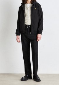 Zwarte ritsvest met capuchon over een lichtgrijze trui, gecombineerd met zwarte broek en zwarte schoenen. Heeft een klein geel logo op het jack.