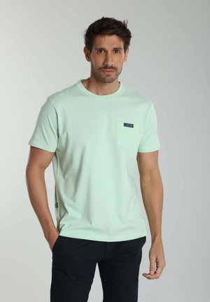Homme portant un t-shirt vert clair à manches courtes avec une poche poitrine et un pantalon noir, debout devant un fond neutre.