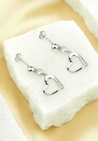 Silver hjärtformade örhängen prydda med en rund kristallpärla och polerade detaljer, uppvisade på en vit sten mot en mjuk, ljus bakgrund.