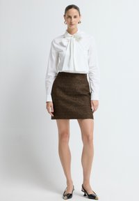 Witte blouse met een grote strikdetail bij de hals, gecombineerd met een korte bruine textuurrok. Zwarte slingback schoenen met gouden accenten.