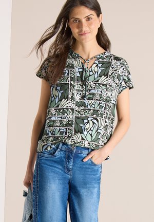 Femme aux longs cheveux bruns portant un chemisier à manches courtes vert à motifs et un jean bleu, une main dans la poche et un léger sourire.