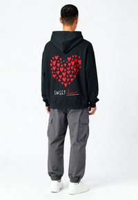 Czarna bluza z kapturem z wzorem w kształcie serca składającym się z czerwonych serc oraz napisem "SWEET heart" na białym tle. Połączona z szarymi spodniami cargo i białymi sneakerami.