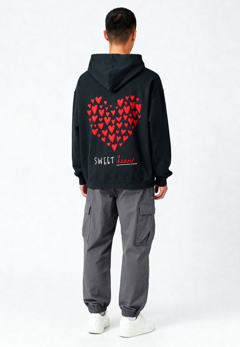 Felpa nera con un design a forma di cuore composto da cuori rossi e la scritta "SWEET heart" in bianco. Abbinata a pantaloni cargo grigi e sneakers bianche.