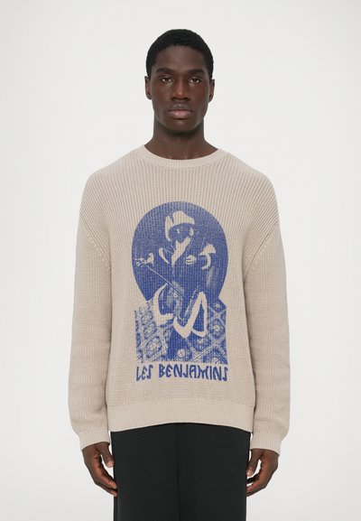 Hombre vestido con un suéter de punto beige con un gráfico azul de una persona sentada y el texto "Les Benjamins", combinado con pantalones negros, frente a un fondo sencillo.