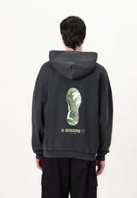 Black hooded sweatshirt with a textured finish. Features a graphic of a green shoe sole on the back with text below. Relaxed fit and ribbed cuffs.