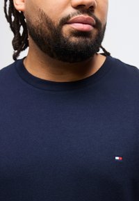 Άτομο με γενειάδα και σκούρο δέρμα που φοράει μπλε πουκάμισο με μικρό ορθογώνιο λογότυπο σε κόκκινο, λευκό και μπλε στο στήθος.