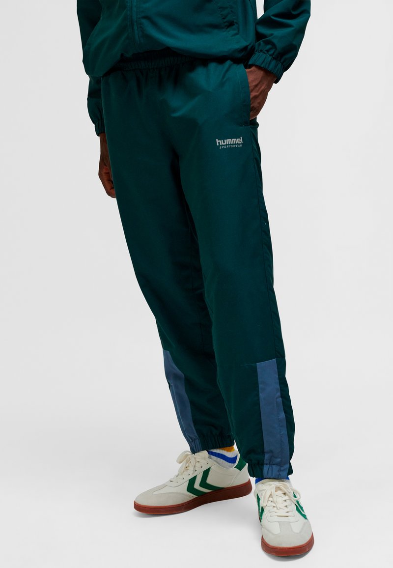 Person trägt dunkelgrüne Hummel-Sporthose mit elastischen Bündchen, mehrfarbige Socken und weiße Turnschuhe mit grünen Chevron-Streifen.