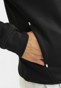 Crna dukserica od mekanog materijala, s bočnom džepom i rebrastim manžetama. Zapaženi bijeli detalj blizu džepa.