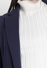 Marinblå kavaj med en fitted, ribbad vit polotröja. Kavajen har en slät textur; polotröjan har vertikala veck för extra detalj.