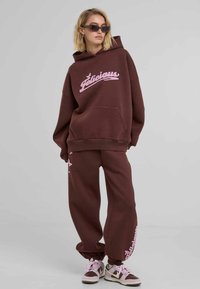 Set de hanorac și pantaloni de trening maro, cu logo-ul brodat roz "Delicious". Include o buzunar frontal și manșete cu cordon; asortat cu adidași maro.