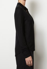 Svart långärmad skjorta med knappar, med en mjuk textur, krage och en lätt kurvatur i fållen. Har knäppning framtill och åtsittande ärmar.