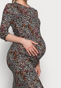 Gravidklänning i mjukt, elastiskt material; har en svart bas med färgglada blommönster i rosa, vitt och grönt. Den figurnära designen framhäver magen.