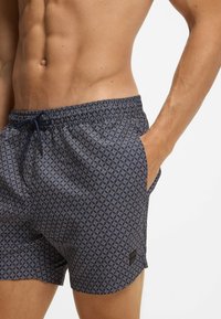 Laivastonsiniset uimashortsit geometrisellä kuvioinnilla, joustavalla vyötäröllä, kiristysnauhalla ja taskuilla. Kevyt kangas, jossa on pehmeä pinta.