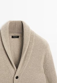 Beige gestrickte Strickjacke mit Schalkragen, die ein strukturiertes Muster aufweist und mit einem einzelnen dunklen Knopf geschlossen wird. Etikett innen im Ausschnitt sichtbar.