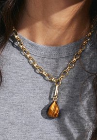 Gold chain necklace featuring large links, with a teardrop-shaped tiger's eye pendant in a polished finish. Worn over a gray shirt.