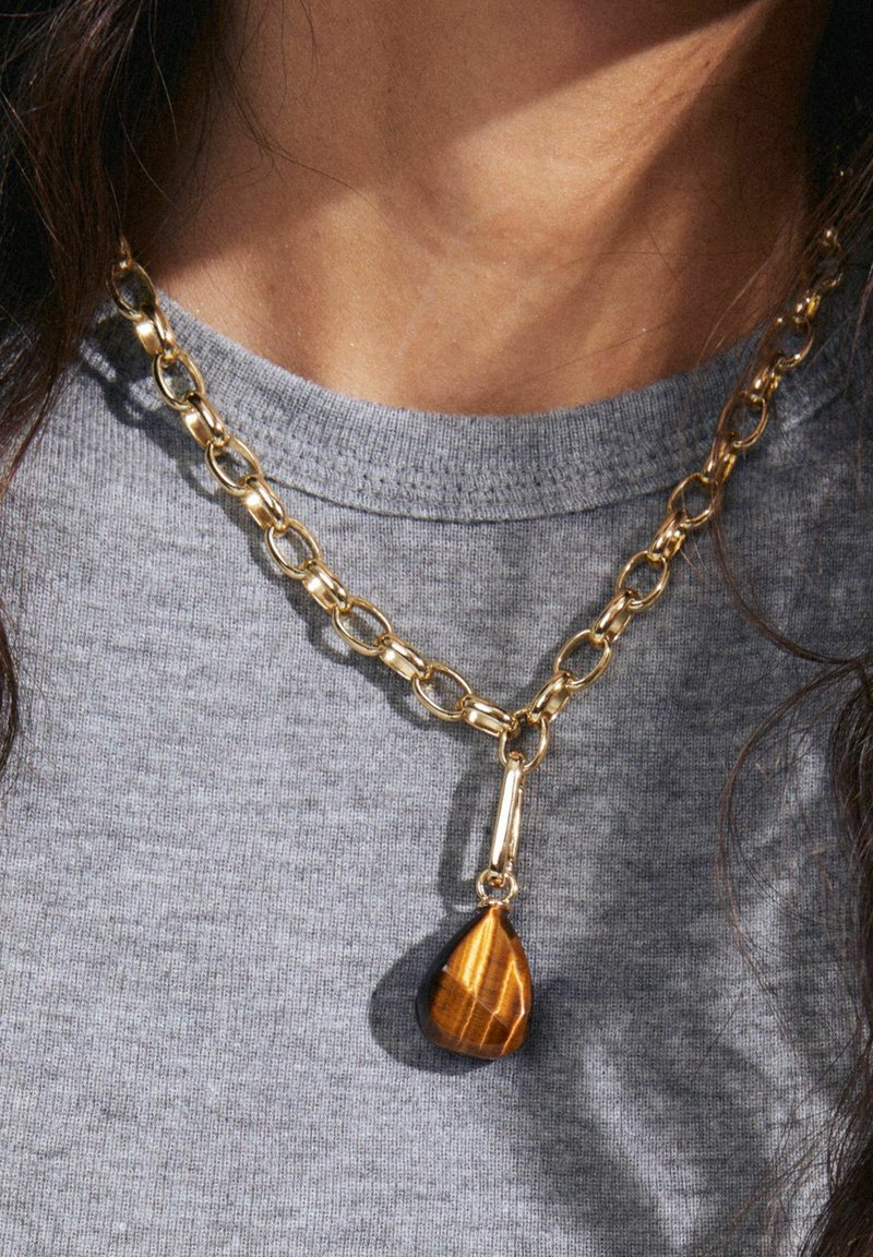 Gold chain necklace featuring large links, with a teardrop-shaped tiger's eye pendant in a polished finish. Worn over a gray shirt.