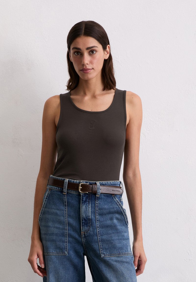 Frau mit schulterlangem dunklem Haar, die ein dunkles, ärmelloses Oberteil trägt, das in eine hoch taillierte blaue Jeans mit braunem Gürtel gesteckt ist, stehend.