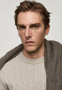 Hellbeige Strickpullover mit Rundhalsausschnitt, kombiniert mit einem dunkelgrauen Wollschal, der über die Schulter drapiert ist. Neutrale Hintergrund.