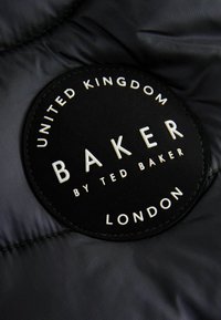 Sort cirkulær label med præget hvid tekst, der står "BAKER by TED BAKER" og "UNITED KINGDOM LONDON" på en quiltet mørk stofbaggrund.