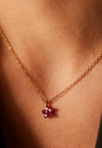 Gouden ketting met een bloemenhanger, versierd met roze edelstenen en kleine witte accenten, op een gestructureerde huidoppervlak.