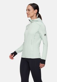 Giacca con cappuccio verde menta chiaro con texture a coste verticali, due tasche frontali con zip e polsini neri. Indossata con leggings neri.