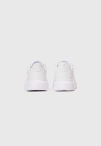 Un paio di sneakers bianche viste da dietro, con suole spesse e un sottile logo in rilievo sulla linguetta del tallone.