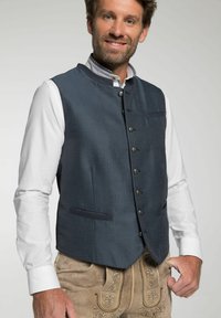 Marineblau gemusterte Weste mit hohem Kragen, Knopfverschluss und zwei Seitentaschen, getragen über einem weißen Hemd und braunen Lederhosen mit Stickerei.