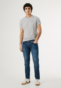 Grijze ronde hals t-shirt, blauwe denim jeans, witte en tan sneakers. Casual pasvorm, gladde textuur, eenvoudig logo detail bij de borst.