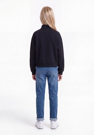 Enfant aux longs cheveux blonds portant un sweat-shirt noir, un jean bleu retroussé et des baskets blanches, debout de dos sur un fond blanc.