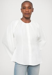 Blusa branca com decote redondo e mangas longas e bufantes. Apresenta um fecho de botões vertical e uma silhueta peplum. Textura suave.