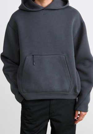 Personne portant un sweat à capuche Nike gris foncé avec une poche avant et un cordon noir, associé à un pantalon noir sur un fond clair uni.