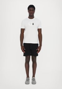 Tricou alb din bumbac cu un imprimeu grafic pe piept, asortat cu pantaloni scurți negri și adidași gri. Modelul stă în fața unui fundal simplu.