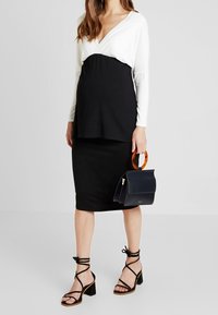 Schwarzes und weißes Langarmoberteil, kombiniert mit einem schwarzen knielangen Rock. Schwarze Riemchenabsätze und eine strukturierte marineblaue Handtasche mit einem Schildpattgriff.