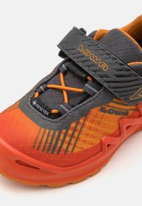 Chaussure de trail en gris et orange, avec une tige en mesh, une étiquette Gore-Tex, des lacets élastiques et une semelle rembourrée avec un motif de traction texturé.