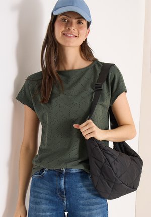 Donna sorridente con berretto blu, maglietta a maniche corte verde oliva traforata, jeans blu, che tiene una borsa a tracolla nera trapuntata.