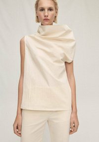 Cream, sleeveless top with a high collar, featuring a draped design that creates texture. Smooth fabric with a slight sheen, paired with neutral pants.