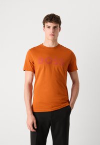 Oransje bomulls t-skjorte med en stor "BOSS"-logo i mørkerødt. Korte ermer, rund hals og en tettsittende fasong. Iført med svarte bukser.