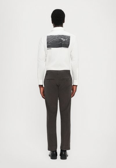 Uomo in piedi, di spalle, con una camicia bianca a maniche lunghe con una stampa océanica bianco e nero sulla schiena, pantaloni scuri e scarpe nere.