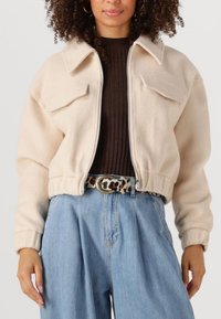 Beige fleecjas met een cropped design, dropped schouders en twee borstzakken. Gecombineerd met lichtblauwe denim met wijde pijpen en een leren riem met luipaardprint.