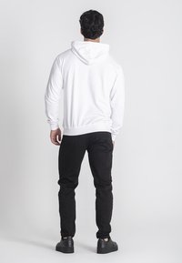 Sudadera blanca con capucha y bolsillo tipo canguro, combinada con jeans negros ajustados y zapatillas negras. Tejido suave, diseño casual.