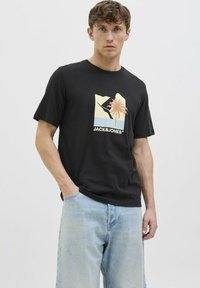 Camiseta de algodón negra con un estampado gráfico de palmeras y paisaje. Combinada con jeans de mezclilla azul claros, el modelo está de pie con las manos en los bolsillos.