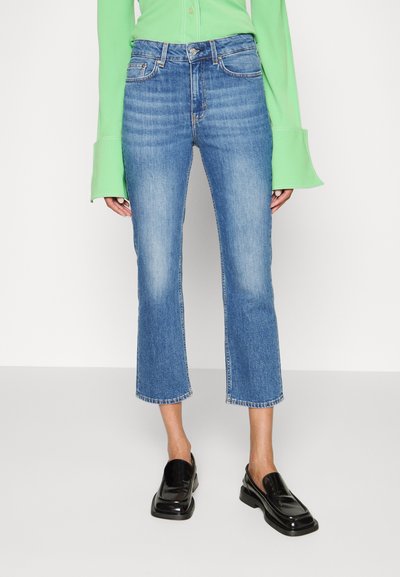 Helle blaue, taillierte Jeans mit geradem Schnitt, fünf Taschen und leicht ausgewaschenem Zustand. Kombiniert mit schwarzen, glänzenden Slippern.