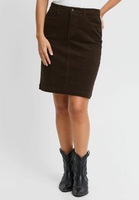Gonna a matita in velluto a coste marrone con tasche frontali, taglio dritto e orlo che si ferma sopra il ginocchio. Indossata con stivali neri in stile cowboy.