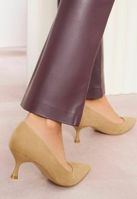Scarpe con tacco alto in suede beige, con punta appuntita e tacco trasparente unico, abbinato a pantaloni skinny in pelle sintetica color prugna.