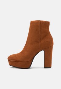 Bottine en suede marron avec un bout arrondi, un haut talon bloc et une semelle plateforme, présentant des lignes épurées et une texture lisse.