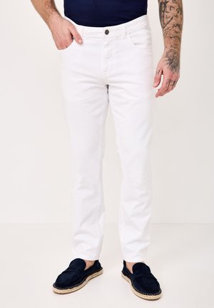 Man wearing slim-fit white jeans, navy blue loafers with woven soles, and a dark blue shirt, hand in pocket, tattooed right forearm visible.