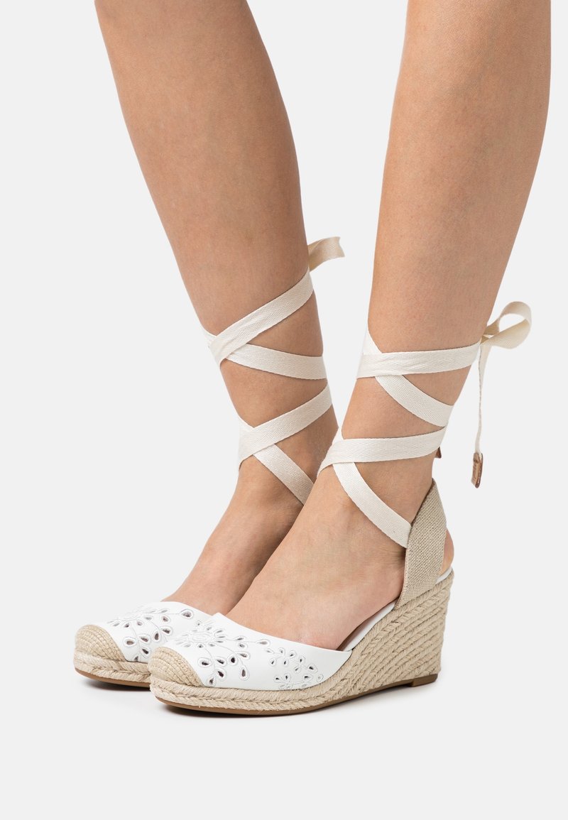 Bílé klínové sandály s výřezovými květinovými detaily, pletenými juty a béžovými kotníkovými pásky obtočenými kolem nohou.