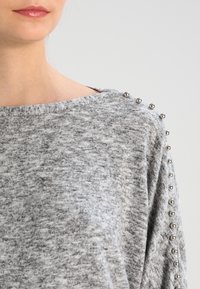 Harmaa neulepaita, jossa on avara kaula-aukko ja metalliset niittikoristeet olkasaumojen varrella. Paita on pehmeää materiaalia ja rentoa mittaa.