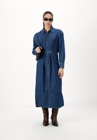 Robe chemise en denim avec des manches longues, une taille ceinturée, un boutonnage sur le devant et des fentes latérales, complétée par des bottes marron à hauteur de genoux et des lunettes de soleil noires.